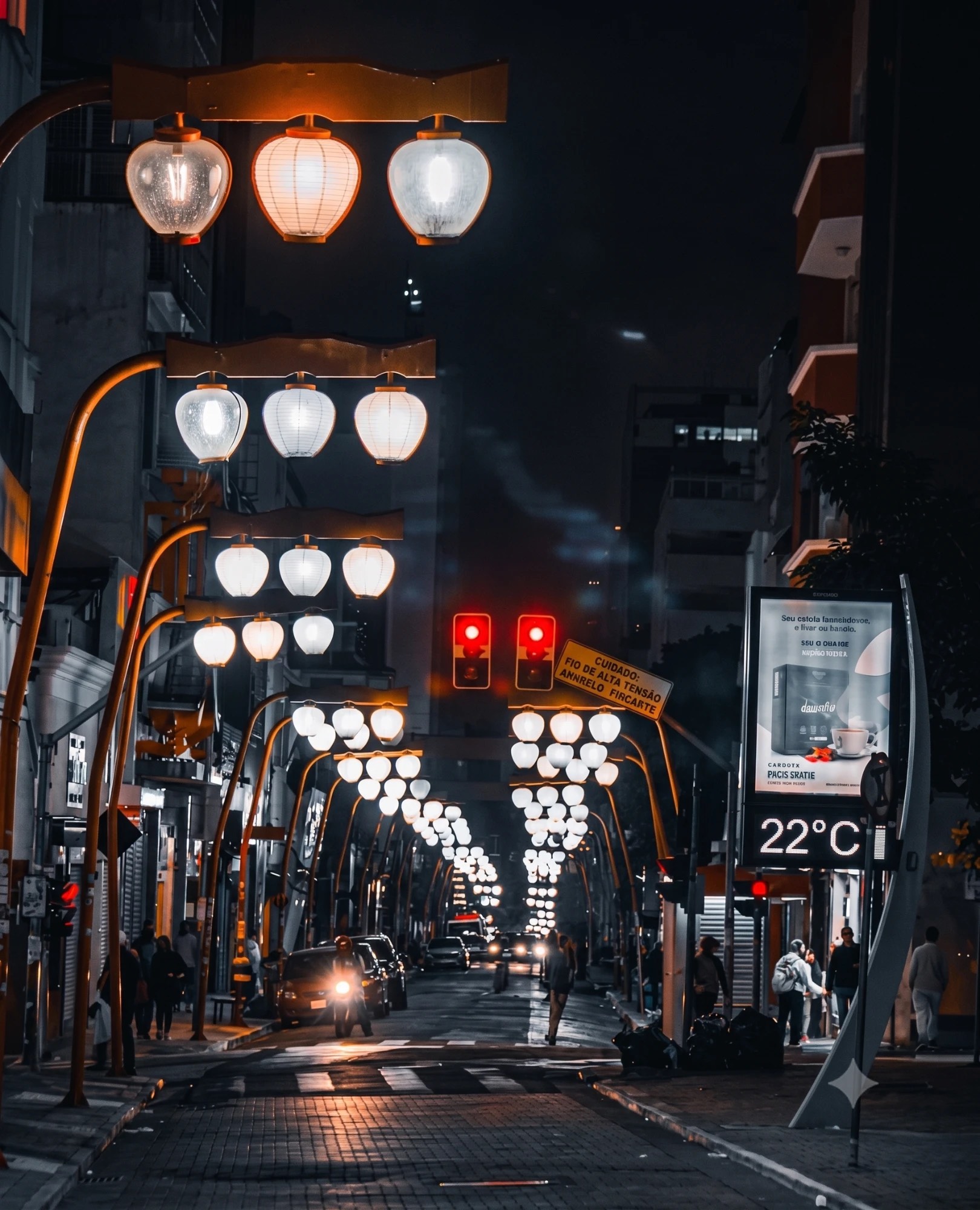 Rua de comércio à noite, iluminada por fileiras de lampiões acesos, com semáforos e pessoas caminhando